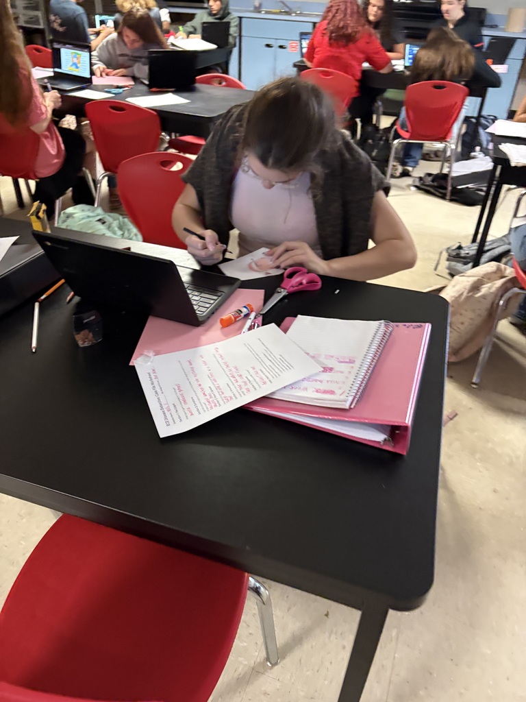 A person sitting at a desk, using a laptop and writing on paper, surrounded by other people.