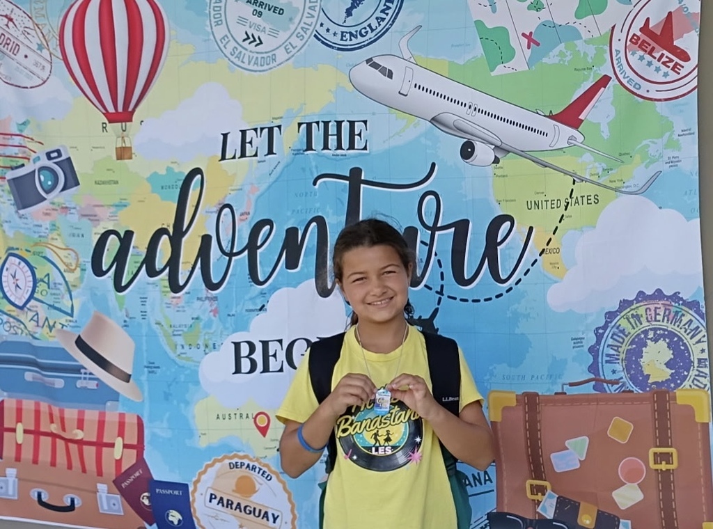 A young girl in a yellow shirt stands before a backdrop with world map, hot air balloon, airplane, and travel gear.
