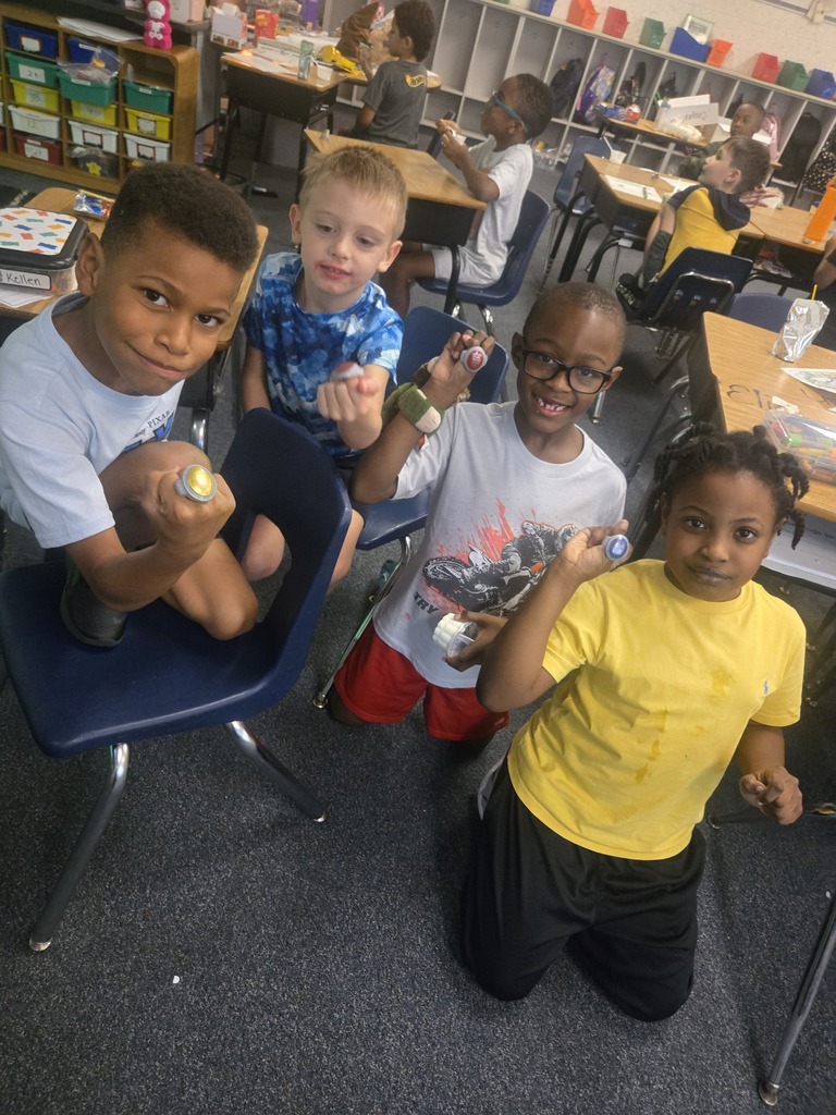 Four children in a classroom, each holding an object, smiling, and posing for the camera.