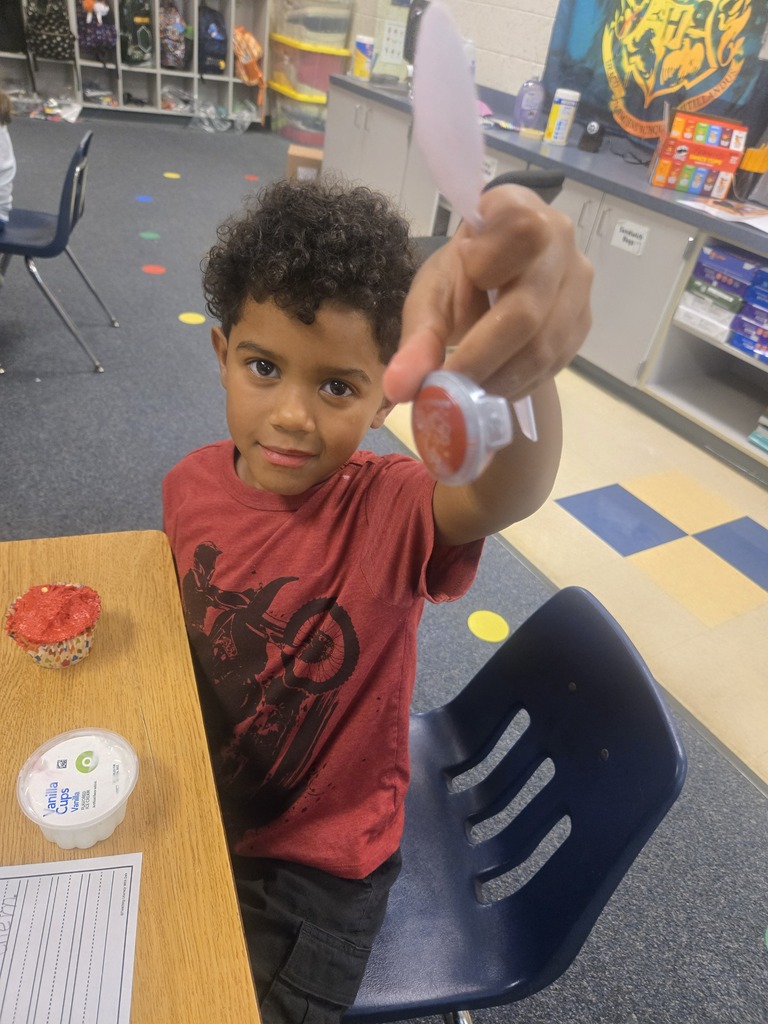 A young child in a red shirt holds up a red object in front of a table.