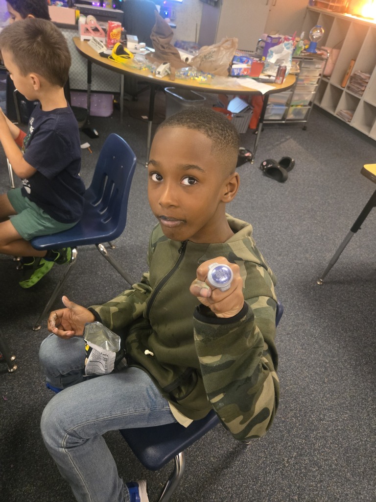 A child sits in a chair holding a small object, smiling, in a classroom with other children.