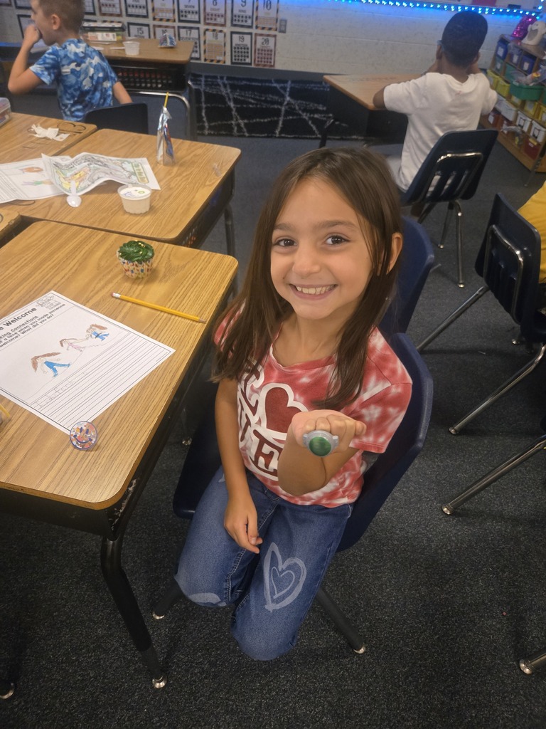 A young girl sits at a desk in a classroom, smiling and posing for a photo.