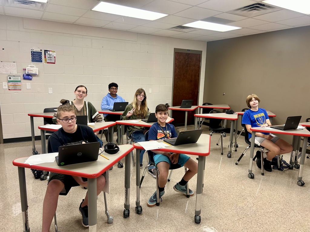 Classroom with desks in rows. Students sit with laptops. Bright lighting. White walls and ceiling. Beige floor.
