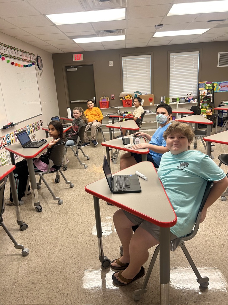 A classroom with students seated at desks, each using laptops. The room has a tiled floor, white walls, and ceiling lights.