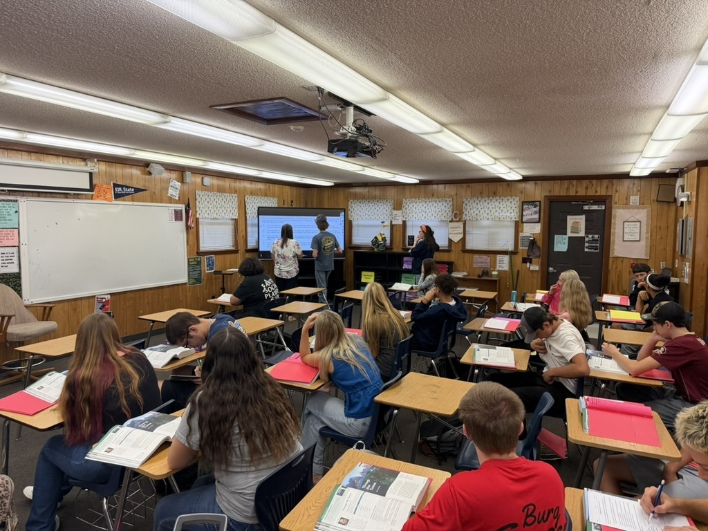 An interior of a classroom with students seated at desks. A teacher stands in front of a screen with a student..
