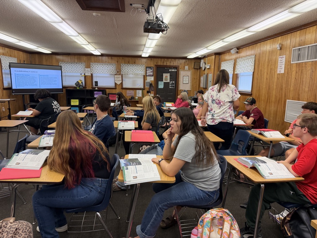 A classroom with wooden walls has students seated at desks. One student works on an assignment.