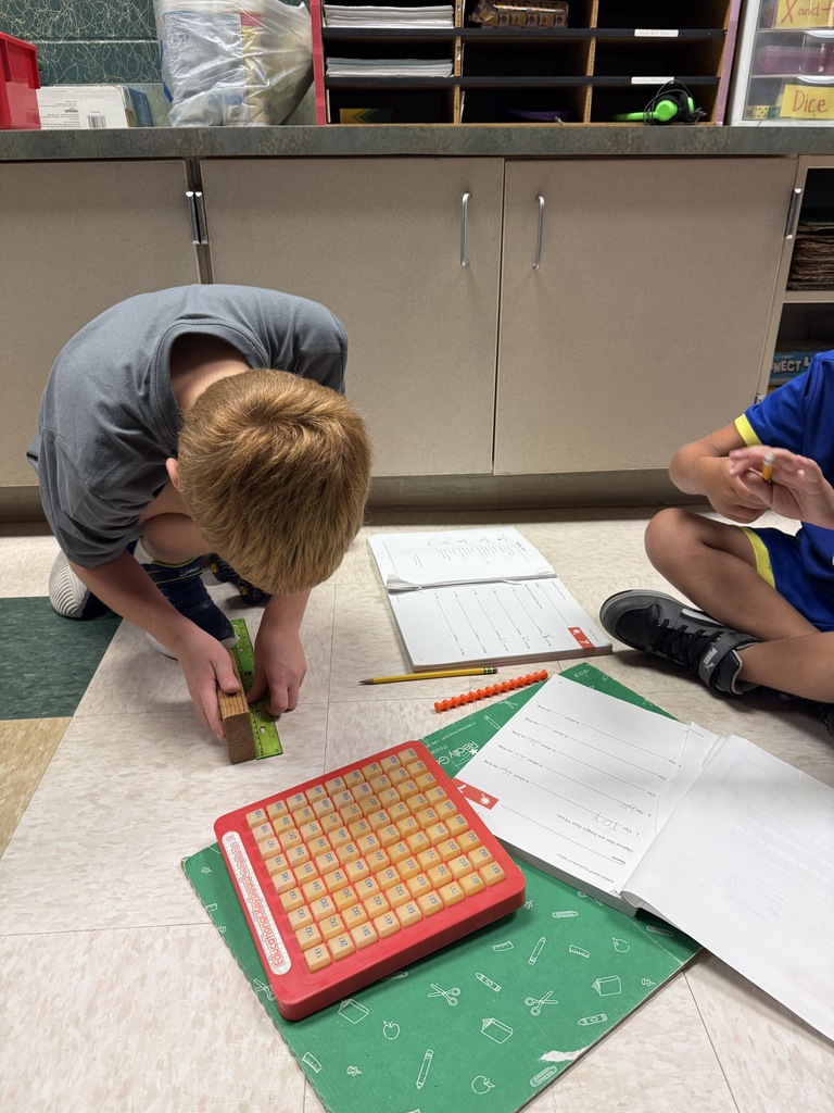 Two children sit on the floor, one examining a red object, the other writing. Papers and a pencil lie on the carpet. A cabinet and storage box are in the background.