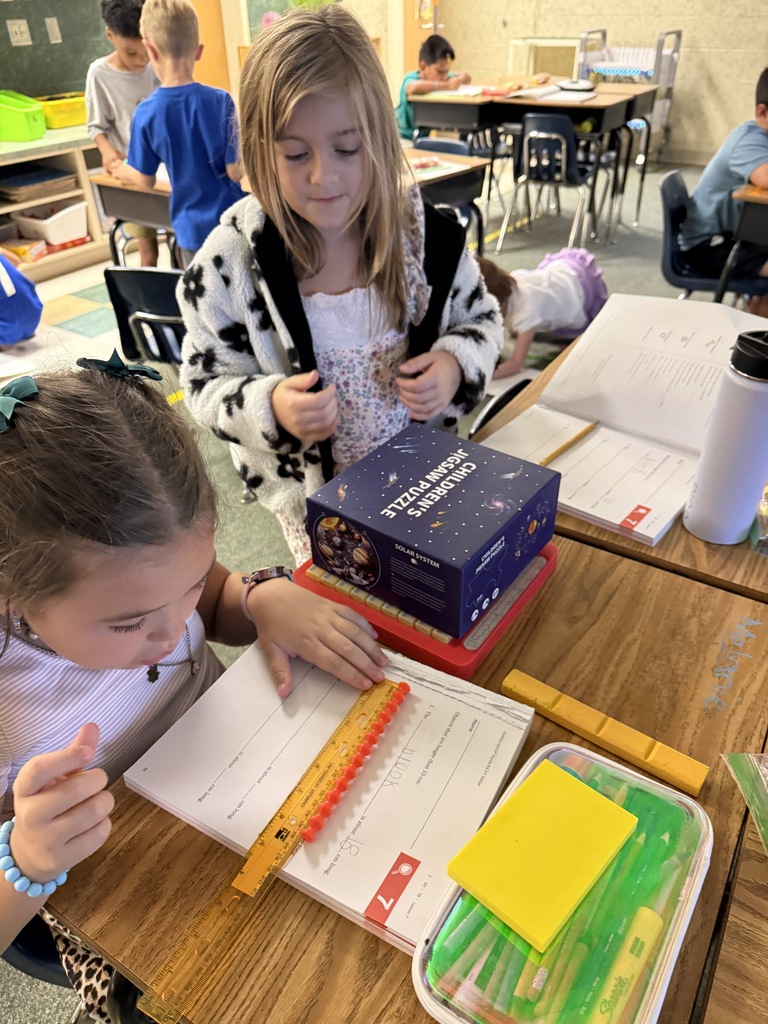 Two young girls, one wearing a bracelet, sit at a table with books, paper, and a ruler.