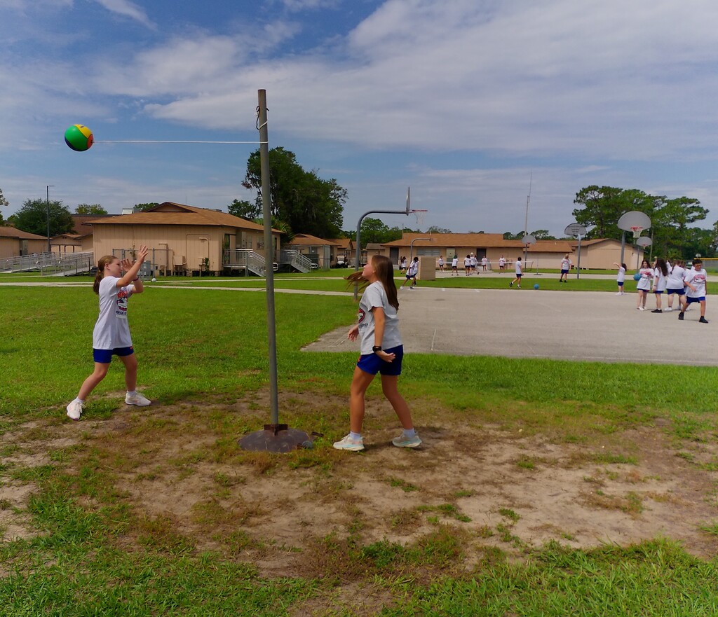 Two girls play volleyball on a field with a net. Other people are in the background near a basketball court.