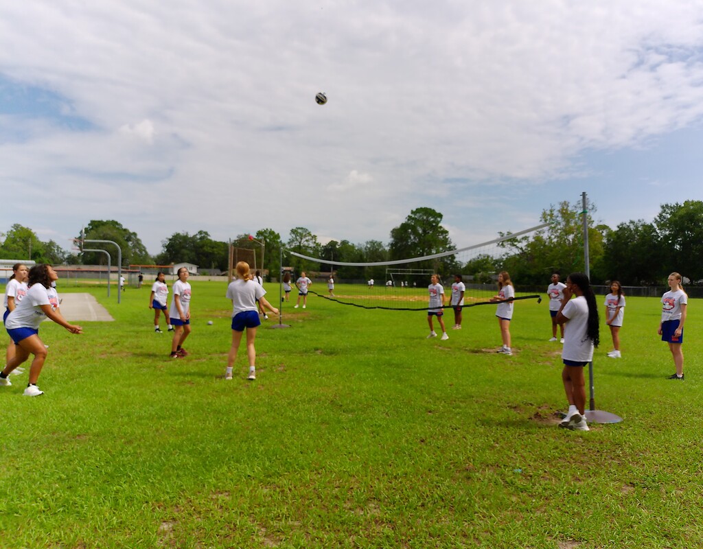 Multiple individuals in white shirts and shorts playing volleyball on a grassy field under partly cloudy skies.