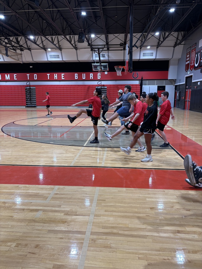 People wearing red shirts, shorts, and sneakers play basketball on an indoor court with red and white walls.