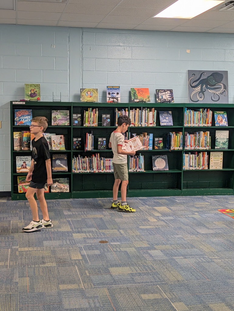 Two children stand in a library, one holding a book, with a bookshelf filled with books behind them.