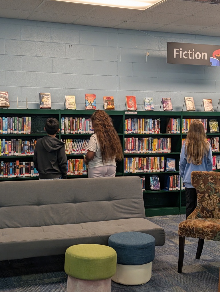 A library with bookshelves labeled "Fiction." A person reads in front of a bookshelf. Two women stand near a chair.