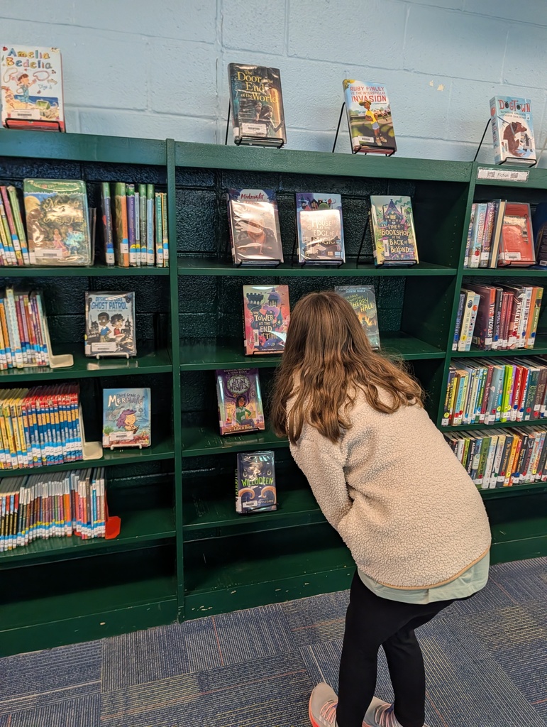 A young girl examines a book while leaning over a bookshelf in a library. The shelves are filled with colorful books.