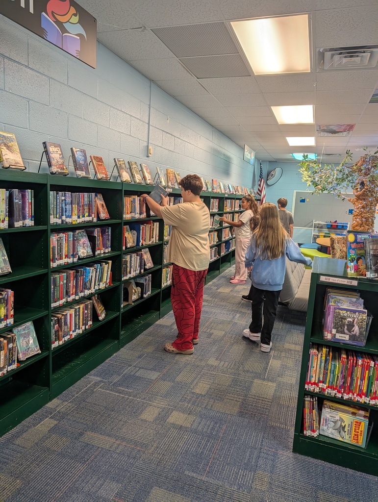 A library with green bookshelves filled with books. People are browsing books on shelves. The ceiling has lights.