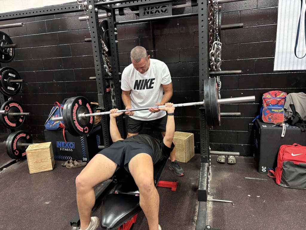 Two men work out in a gym. One man spots another man lifting a barbell while lying on a bench.