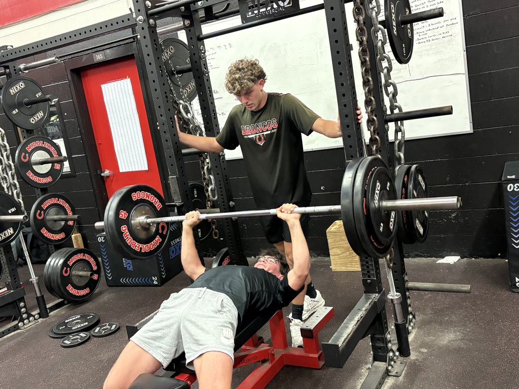 Two men work out in a gym. One man spots another man lifting a barbell while lying on a bench.