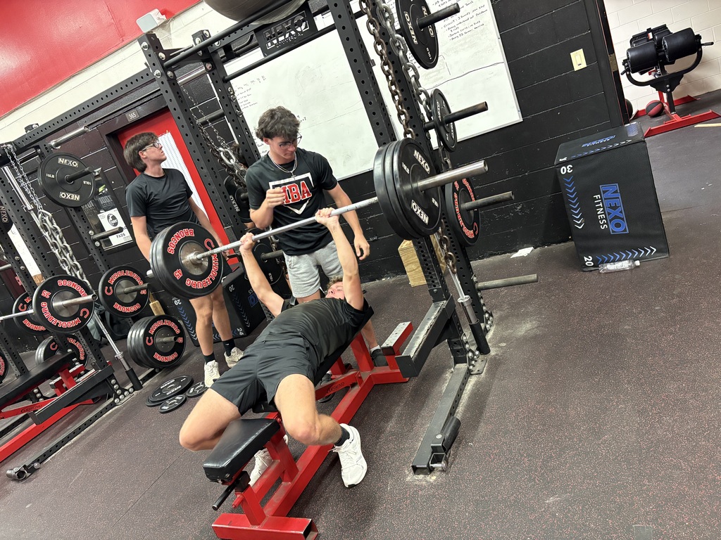 Three people are in a gym. One is performing a bench press with assistance from two others.