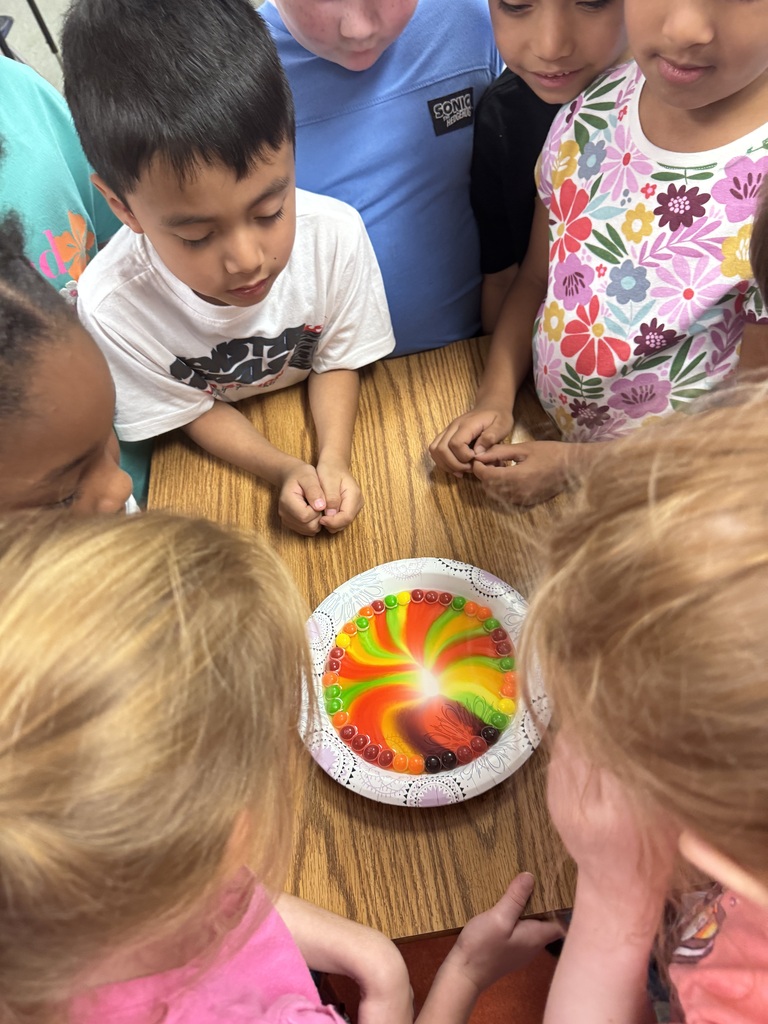 Children gather around a table with a colorful plate on it. One child is wearing a white shirt.