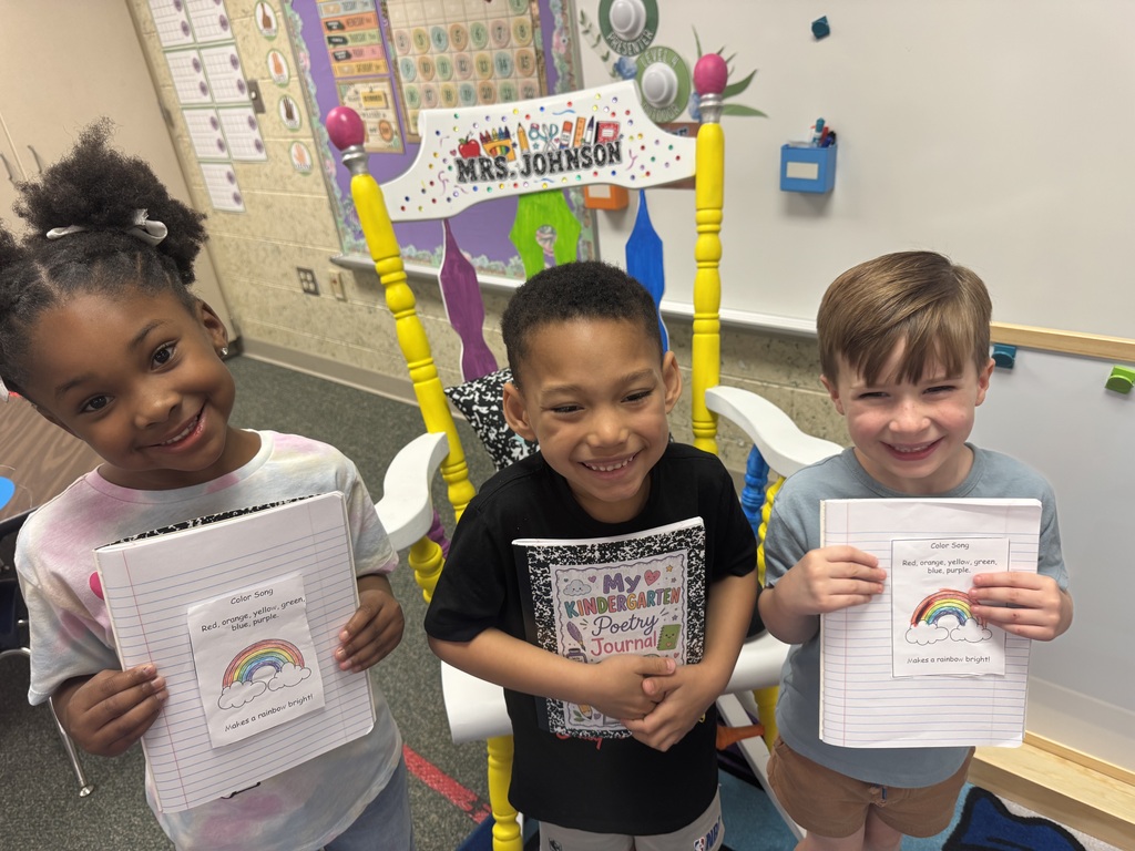 Three children stand behind a chair holding up papers. One holds a rainbow-themed paper. Behind them is a white board.