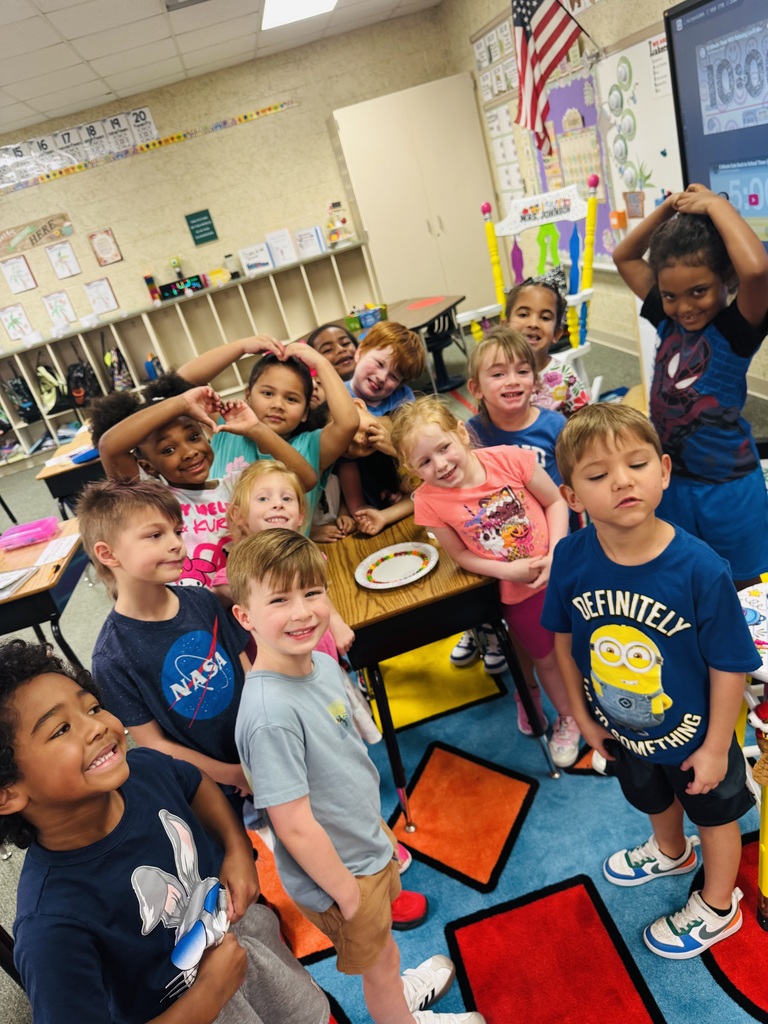 Kids in a classroom pose for a group photo. Some stand while others sit. A teacher is present.