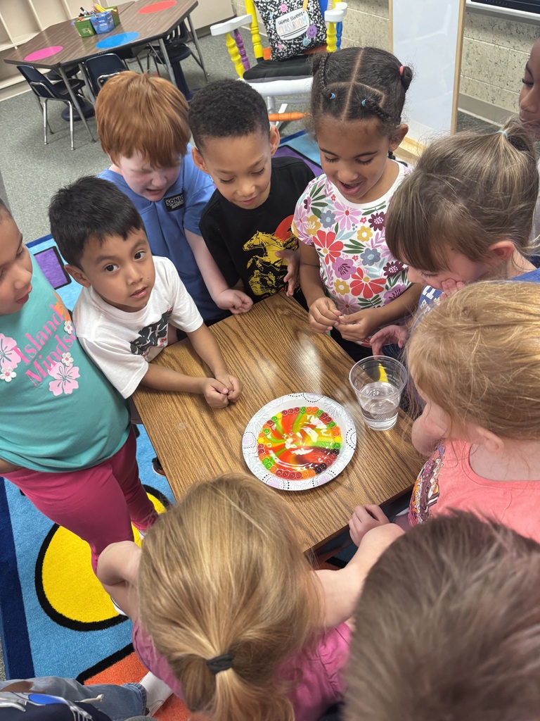 Several children gather around a table to observe a colorful plate of food. They are in a classroom with chairs and desks.