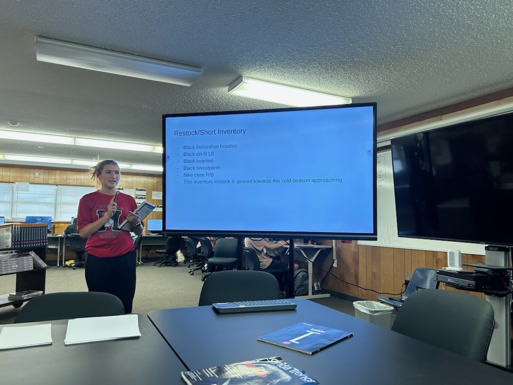 Woman in a red shirt stands in front of a large screen showing text. A table with books and a remote sits in front of her.