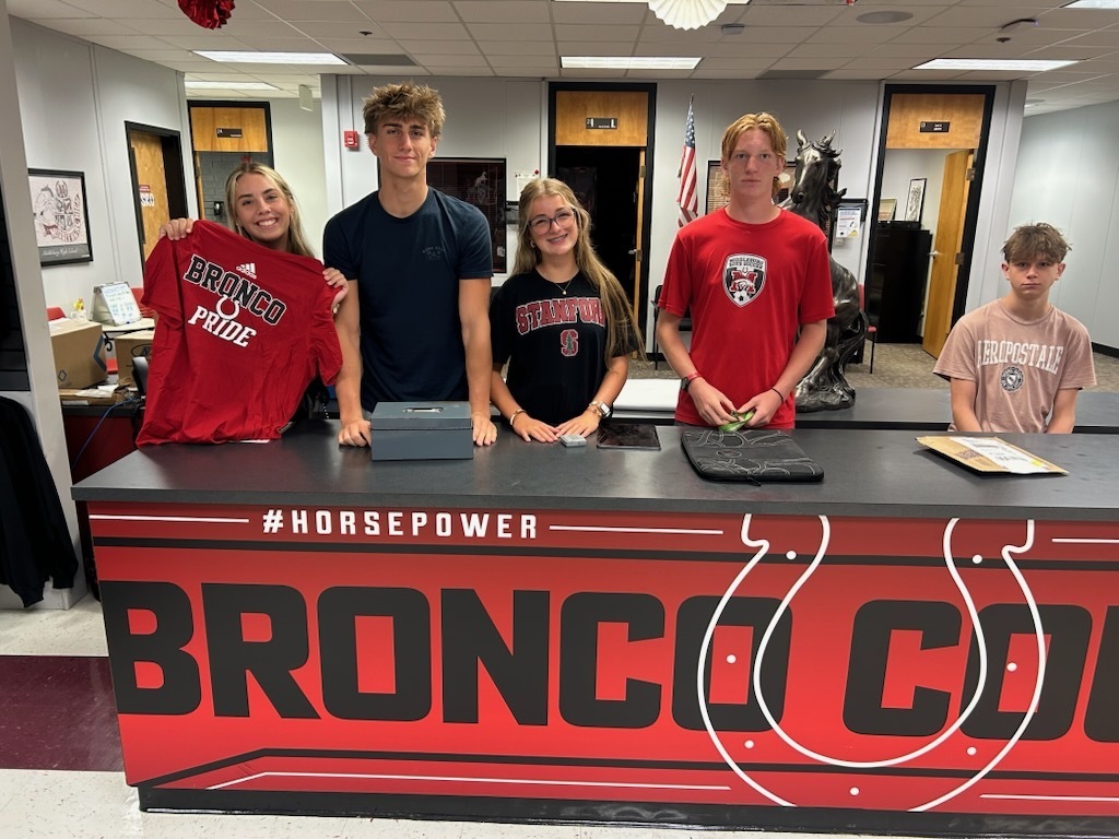 Five young people stand behind a red desk, holding red shirts, with one holding a sign that reads "BRONG POWER".