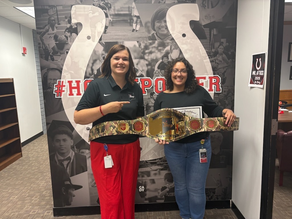 Two women stand in front of a large poster with a horse shoe and text. One holds a belt.