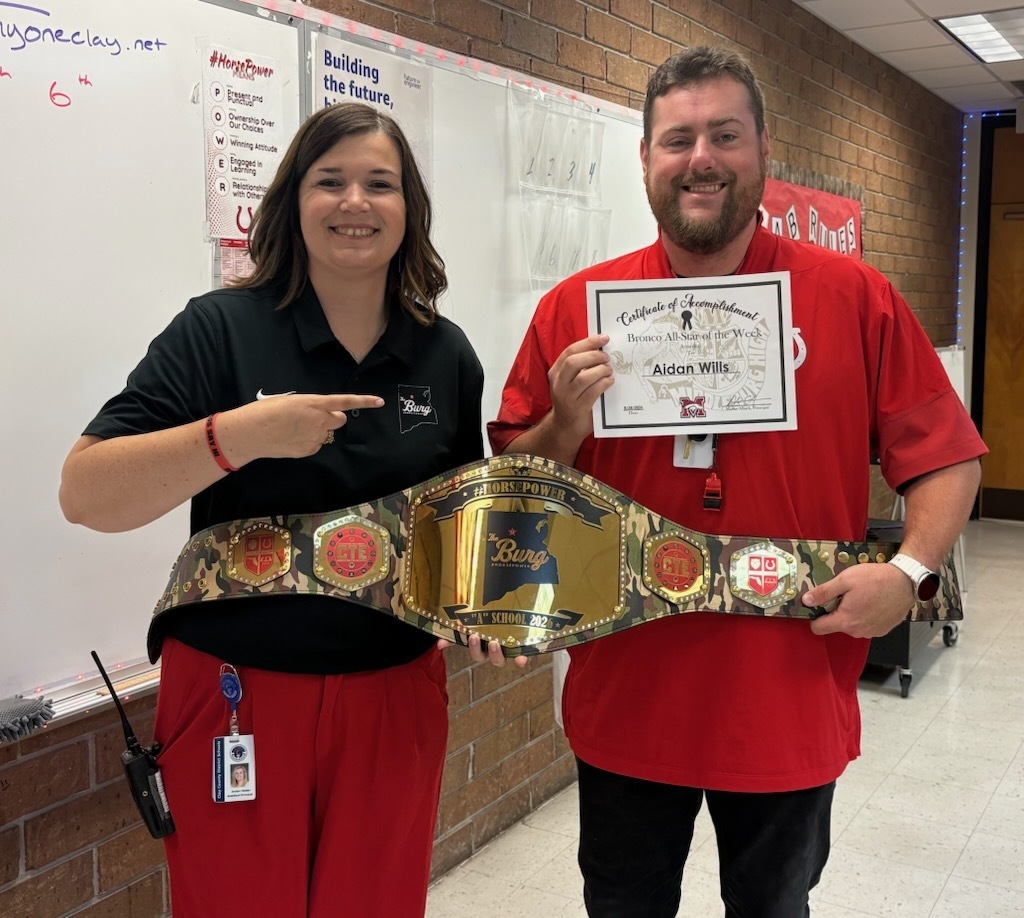 A man and woman hold a championship belt. The man holds a certificate. They stand in a classroom. A whiteboard is behind them.