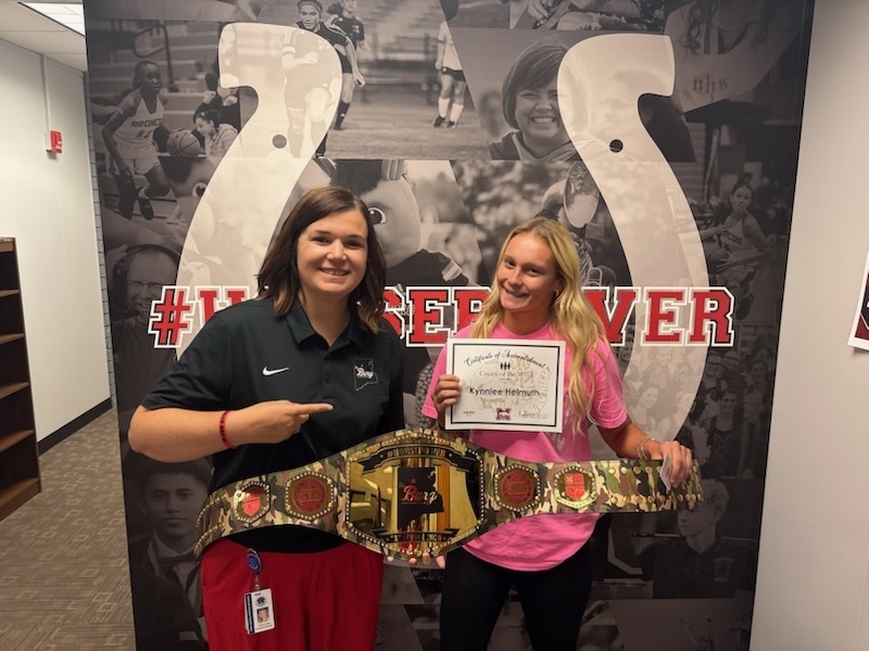 Two women pose in front of a large photo collage. One holds a certificate, the other a belt.