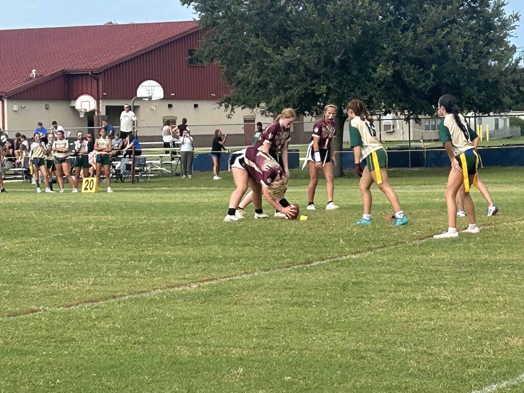 A group of players in jerseys playing a game on a field. A player is bending down. Spectators are watching from bleachers.