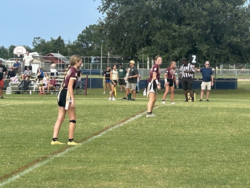 Field with a white line. Players stand in maroon uniforms. A referee holds a number sign. People watch from bleachers.