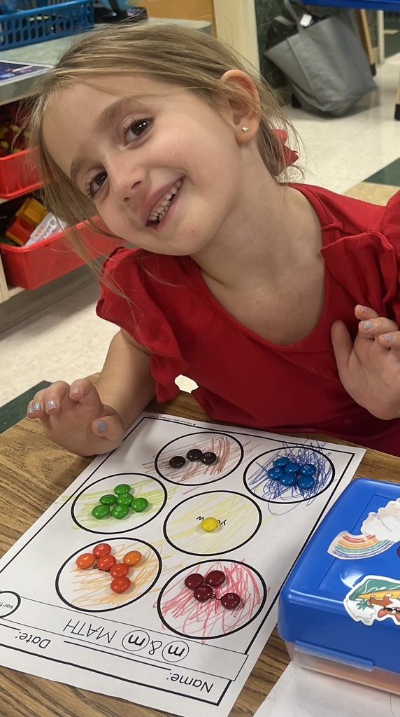 A young girl sits at a table with a worksheet showing circles filled with different colored marbles. She smiles and looks at the camera.