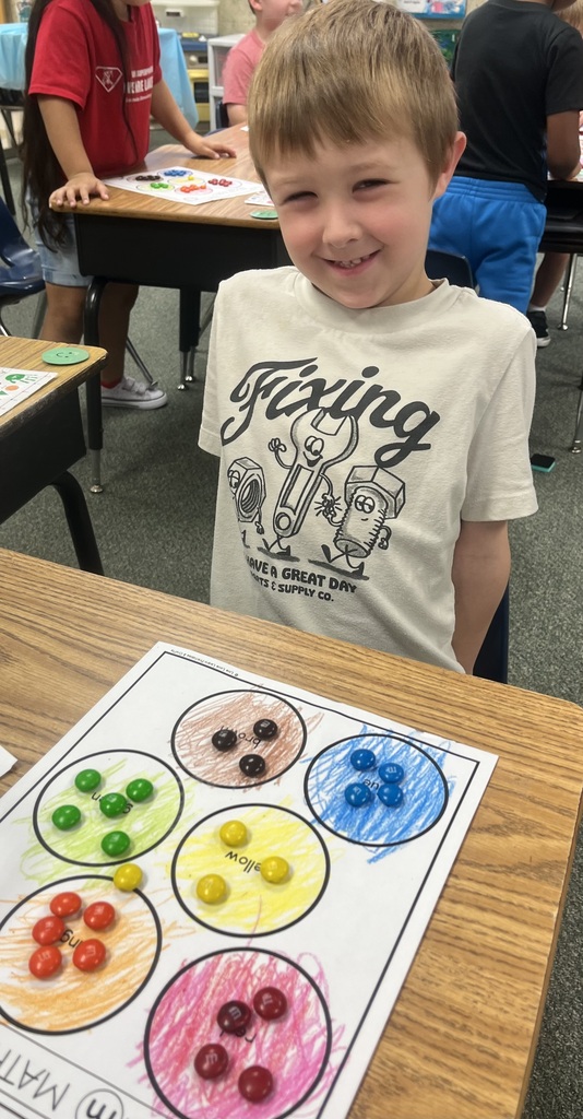 A young boy sits at a table with a coloring worksheet featuring circles with candies. He smiles at the camera. Behind him, two more children are working at desks.