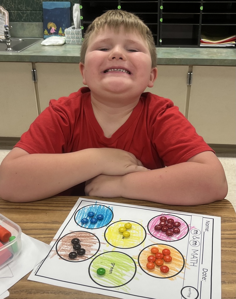 Child seated at a table with arms folded, smiling, wearing a red shirt. In front of him, a colorful activity sheet with various colored beads on it.