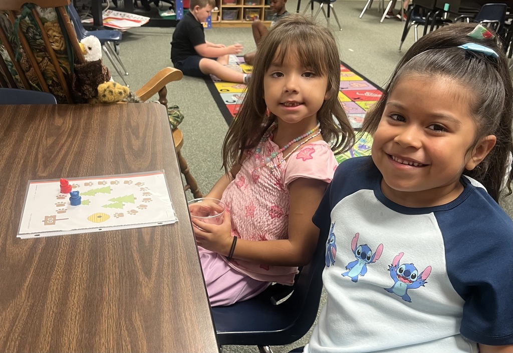 Two young girls sitting at a table with a game board, one wearing a pink shirt.