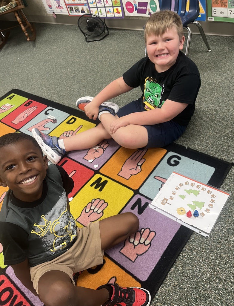 Two children sit on a colorful mat with alphabet letters and hands drawn on it. One smiles and the other sits next to a book.