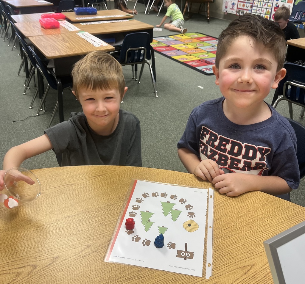Two boys in a classroom, seated at a table. One holds a glass with a liquid, and the other smiles at the camera.