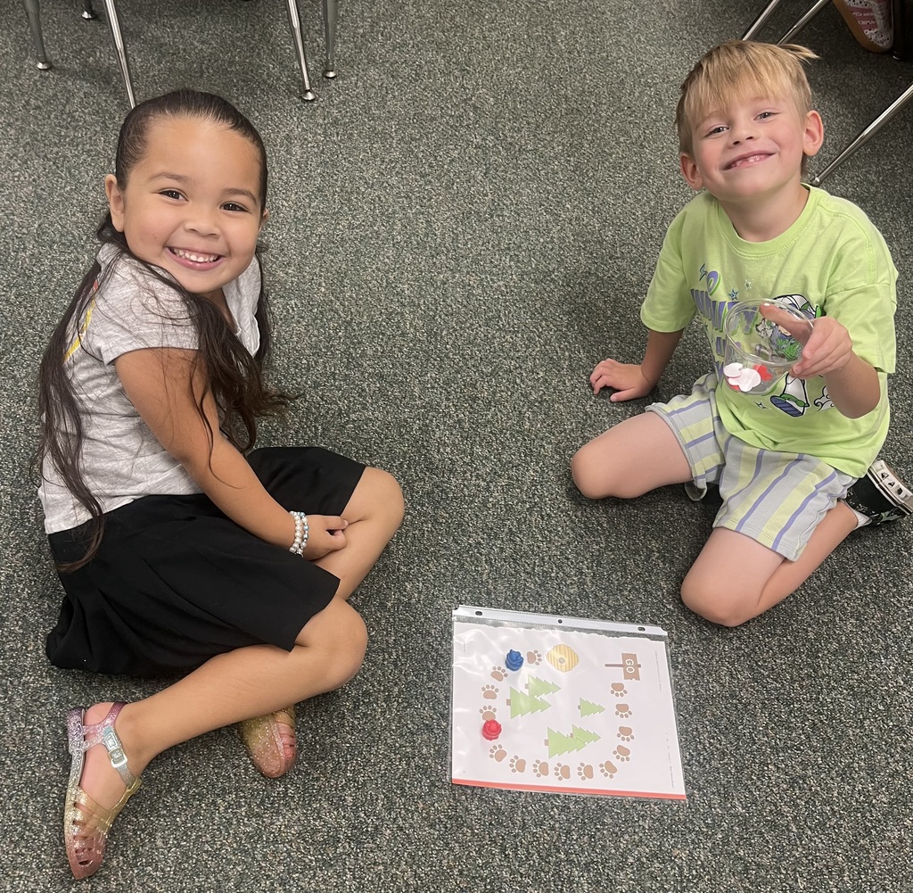 Two children sit on the floor, smiling and facing each other. One holds a colorful page with a spiral design.