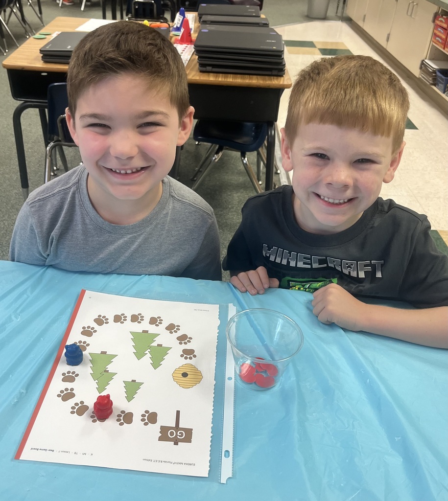 Two young boys smile and sit at a table. One holds a board with a Christmas tree and colored dots.