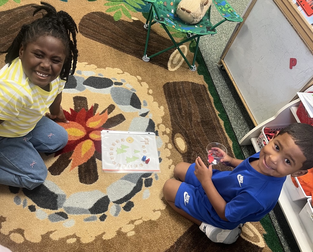 Two children sit on a rug with a campfire design, one holds a small container and the other smiles.