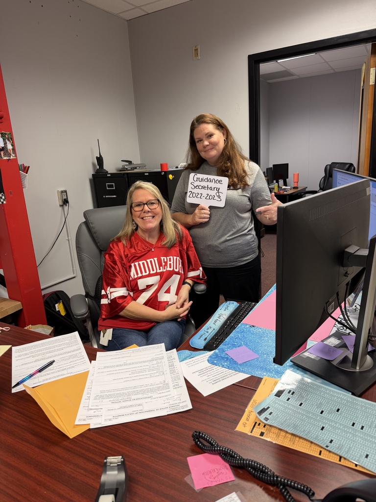 Two people in an office. One holds a sign. A computer, desk with papers, pens, and office supplies.