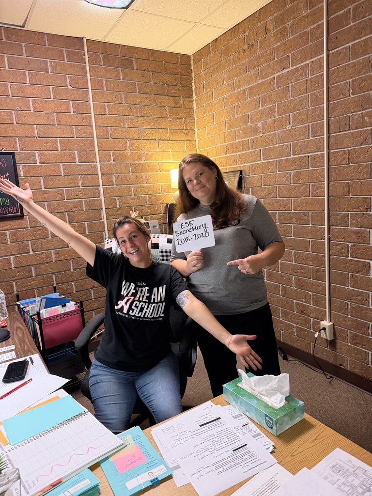 Two people pose in a room with brick walls, one holding a sign. Papers, notebooks, and tissues are on the desk.