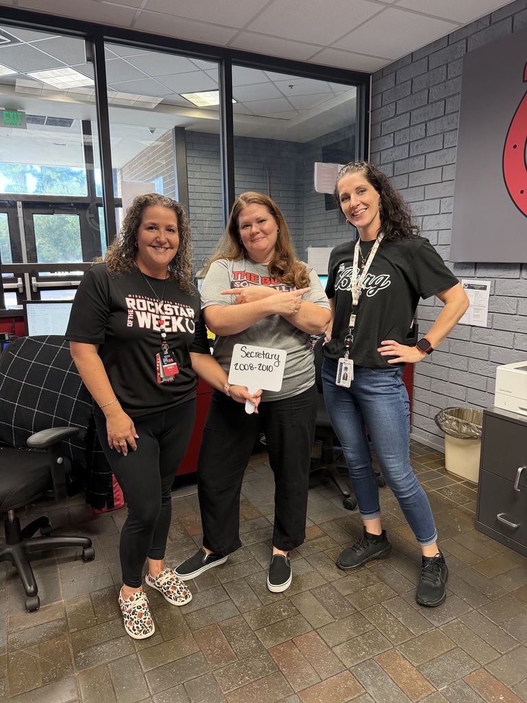 Three women in black shirts and jeans standing in an office room with brick walls. One holds a sign.