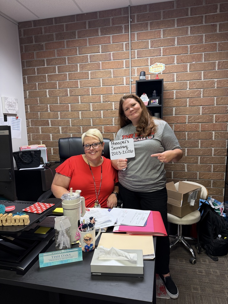 Two women stand behind a desk with various office supplies. One wears glasses and a red shirt; the other, a gray shirt. The background is a brick wall.