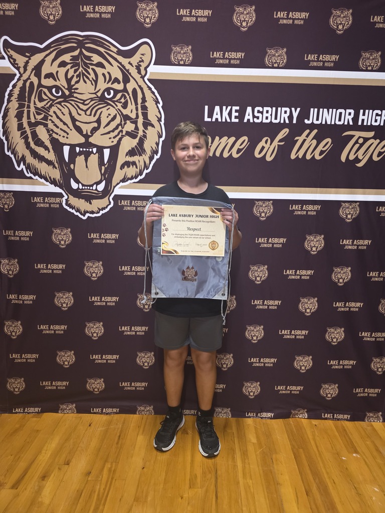 A person stands on a wooden floor, holding a certificate, and smiling in front of a wall with a tiger logo.