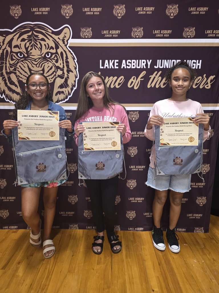 Three students pose with certificates in their jeans vests, standing on a wooden floor in front of a Lake Asbury Junior High banner.