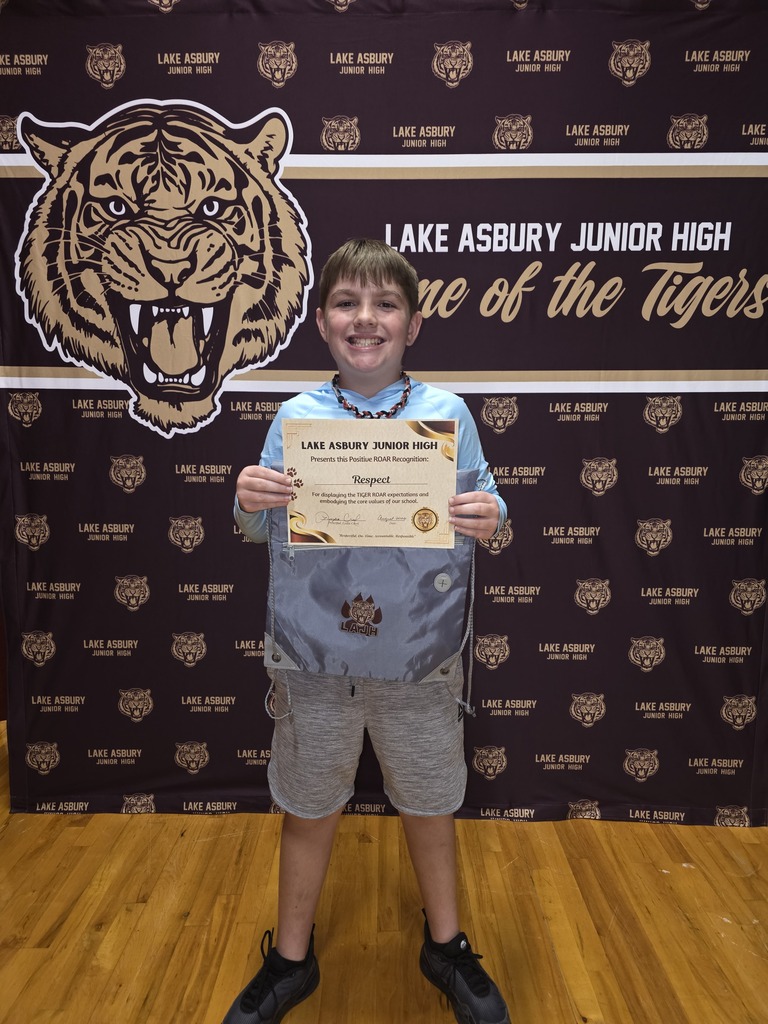 A young boy stands in front of a backdrop with a tiger logo. He holds a certificate and wears a blue shirt, shorts, and sneakers.