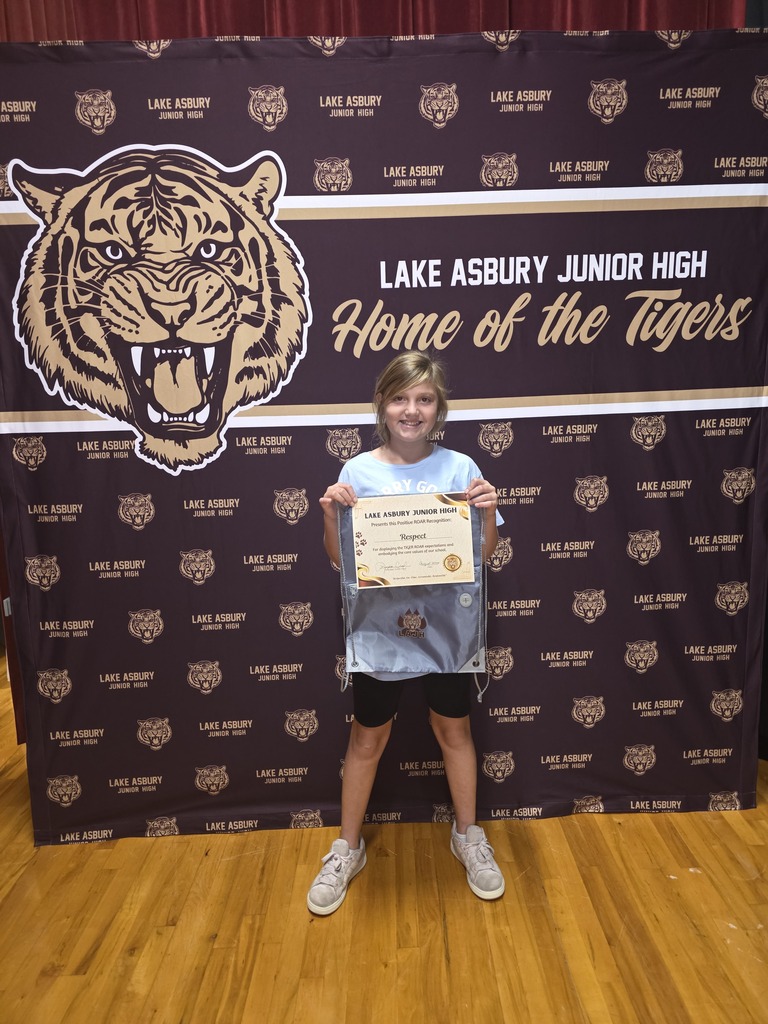 A young child stands on a wooden floor, holding a certificate. Behind them is a backdrop with a tiger logo and text reading "Lake Asbury Junior High Home of the Tigers."